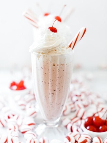 Chick Fil A peppermint milk shake in a tall glass topped with whipped cream, a cherry, and a peppermint stick. There are peppermint candies scattered around it.
