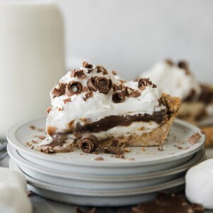 Slice of Smores pie on a stack of plates with a cream cheese toasted marshmallow later, chocolate custard layer and generous whipped cream.