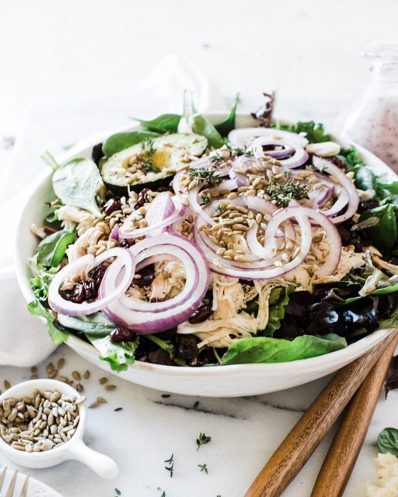 A ¾ view of turkey salad topped with red onions. There are salad servers to the side and a bottle of dressing in the background.