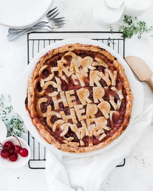 Homemade cherry pie in a white pie dish. The dish is set upon a wire cooling rack and is surrounded by fresh time, sugar, and cherries.