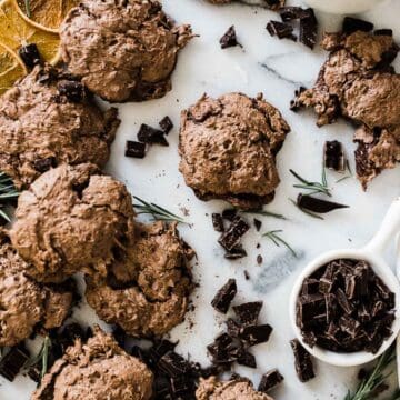chocolate meringue cookies scattered across a marble board. There are chunks of chocolate, rosemary, and dried oranges scattered around.