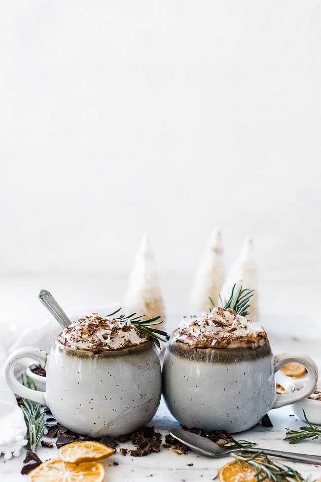 Two mugs filled with coconut hot cocoa. There are bottle brush trees in the background.