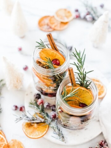 Stovetop Potpourri in glass mason jars. There are bottle brushes, rosemary, cranberries, and orange slices surrounding.