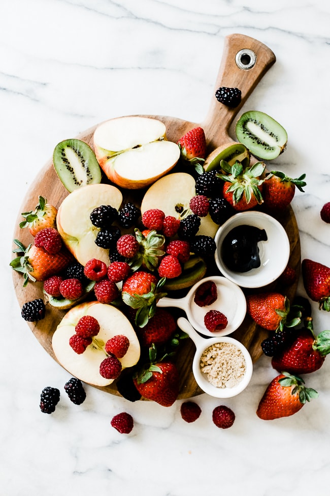 Apples, kiwi, strawberry, blackberries, and raspberries scattered on a wooded cutting board.
