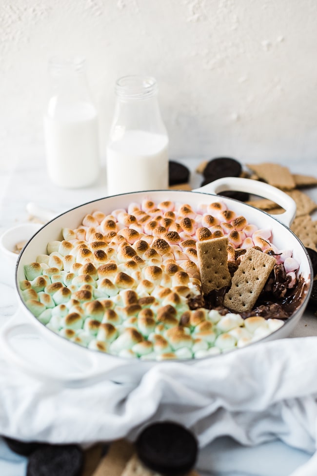 A ¾ view of S'mores dip recipe in a white braiser. There are bottles of milk in the background and the braiser is surrounded by graham crackers.