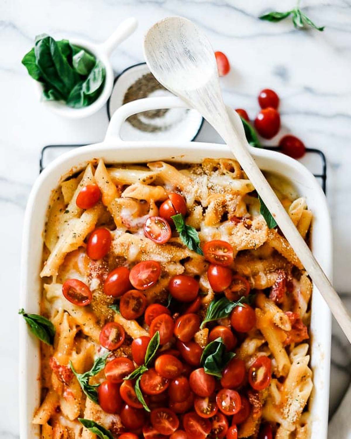 An overhead shot of half a pan of penne pasta bake in a white roasting pan. There is a small bowl of basil and a small bowl of pepper to the side.