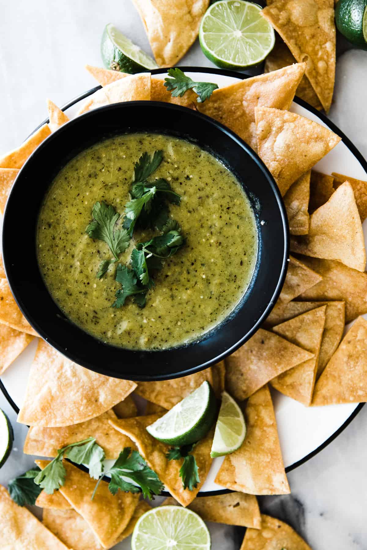 Tomatillo salsa verde in black bowl on a plate surrounded by tortilla chips.