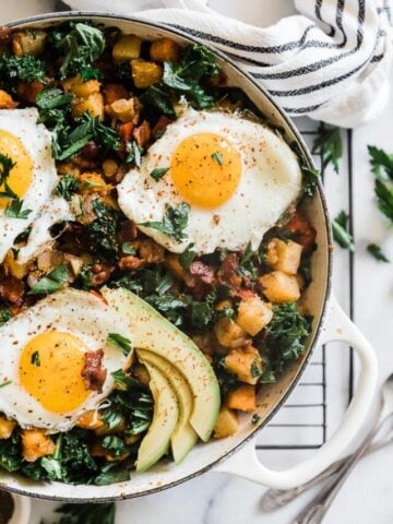 A close up of potato hash recipe in a white braiser. There are sunny Side up eggs on a top and parsley.