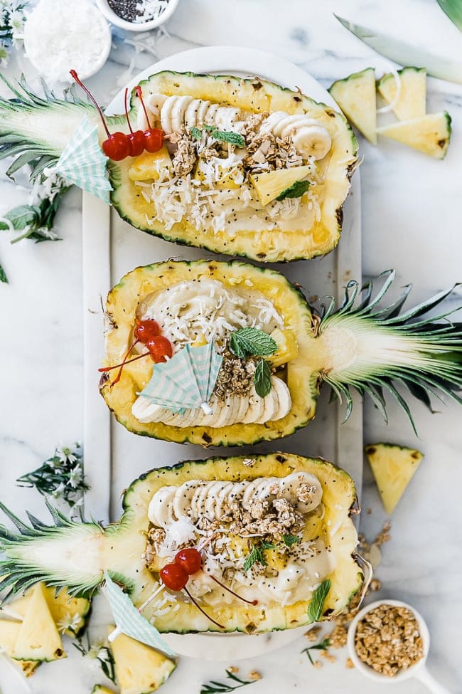 Three pineapple halves on a marble tray filled with Pina colada smoothie and toppings. There are small bowls of granola, coconut, and chia to the side.