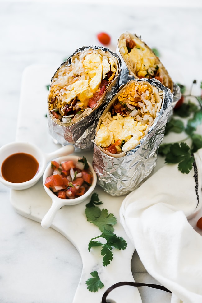 a ¾ shot of 2 breakfast burritos wrapped in foil. They are set beside cilantro and a small bowl of pico.
