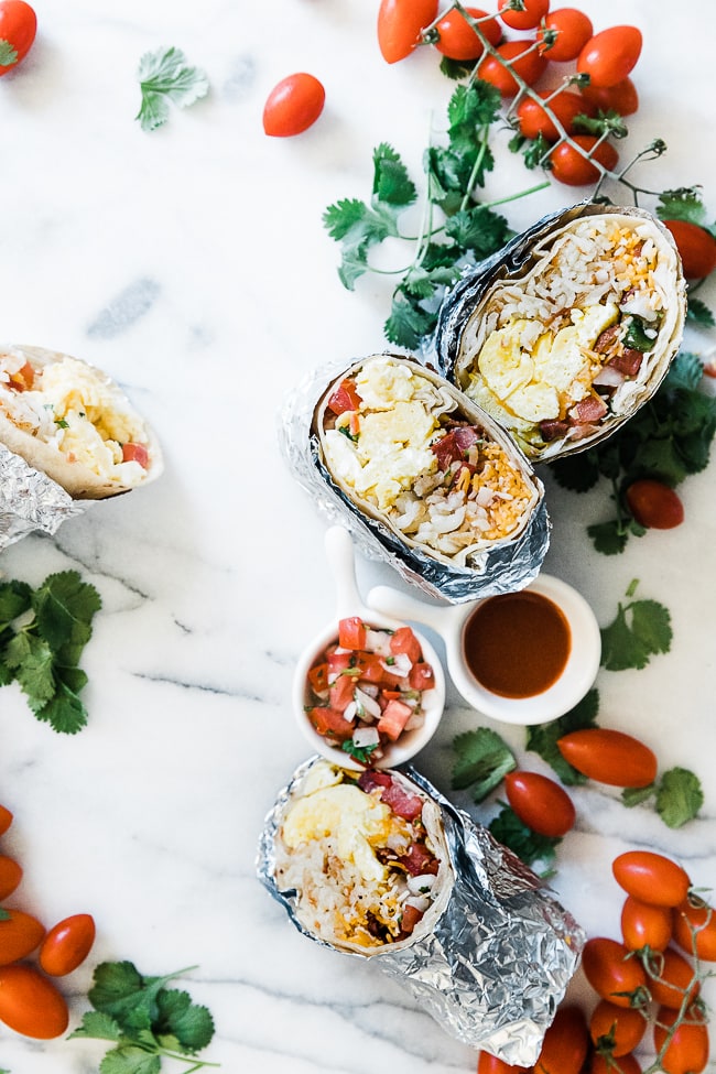 An overhead shot of breakfast burritos scattered on a marble counter surrounded by tomatoes and cilantro.