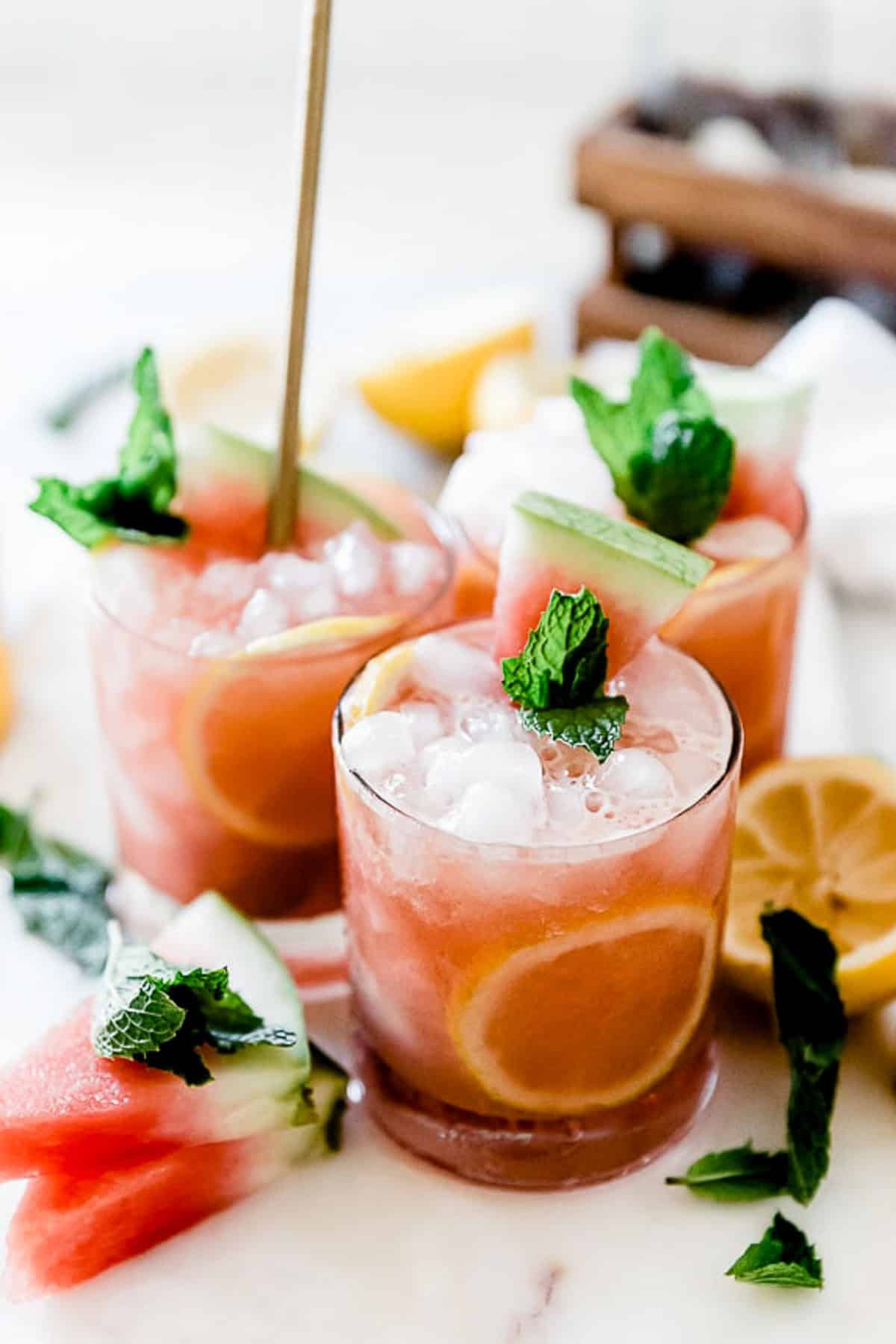 Three glasses of watermelon lemonade set together on a marble tray, surrounded by watermelon slices and lemons.