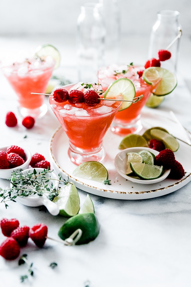 Homemade raspberry limeade in martini glasses, set onto of a white tray surrounded by limes and raspberries.
