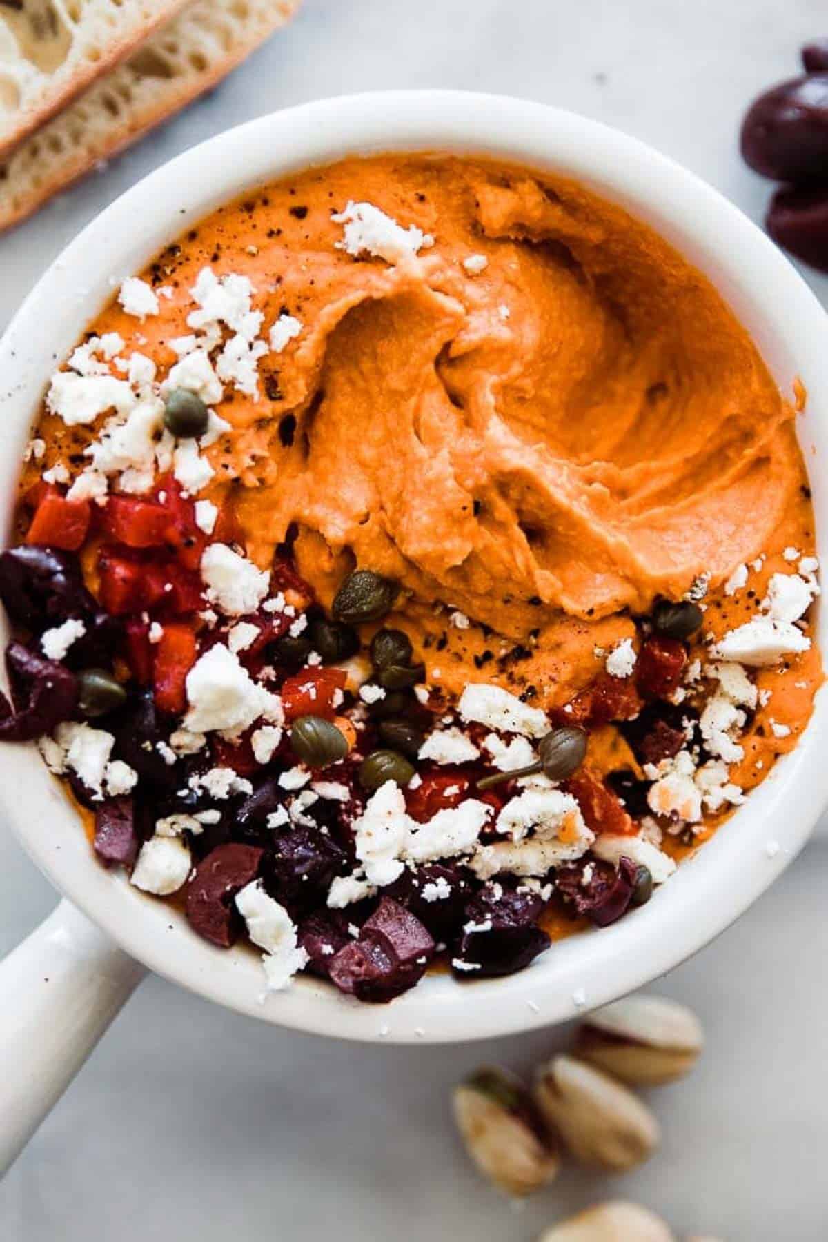 A close up of roasted red pepper hummus topped with feta, olives, and peppers.