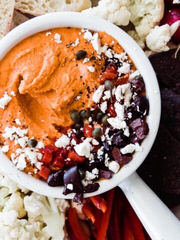 Roasted red pepper hummus in a white bowl surrounded by veggies and bread.