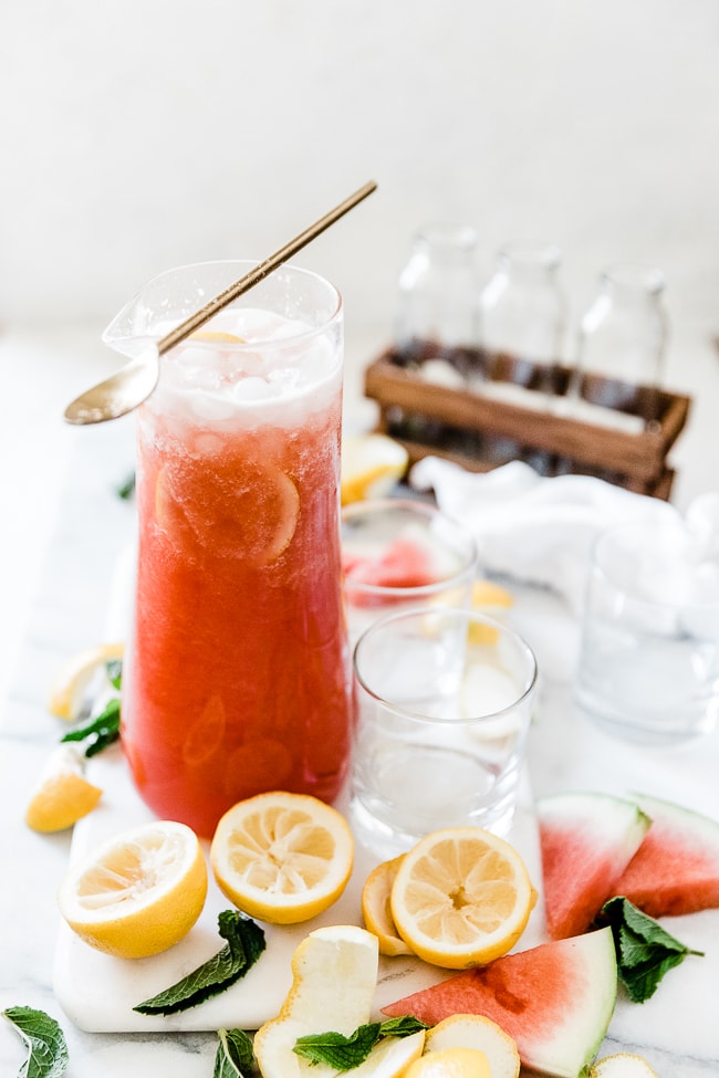 Watermelon lemonade in a tall pitcher surrounded by glasses and lemon sliced.