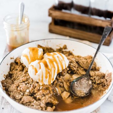 Caramel apple crisp in a white braiser, topped with ice cream. There are glass bottles in the background.