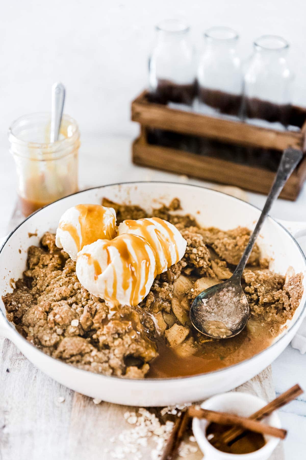 Caramel apple crisp in a white braiser, topped with ice cream. There are glass bottles in the background.