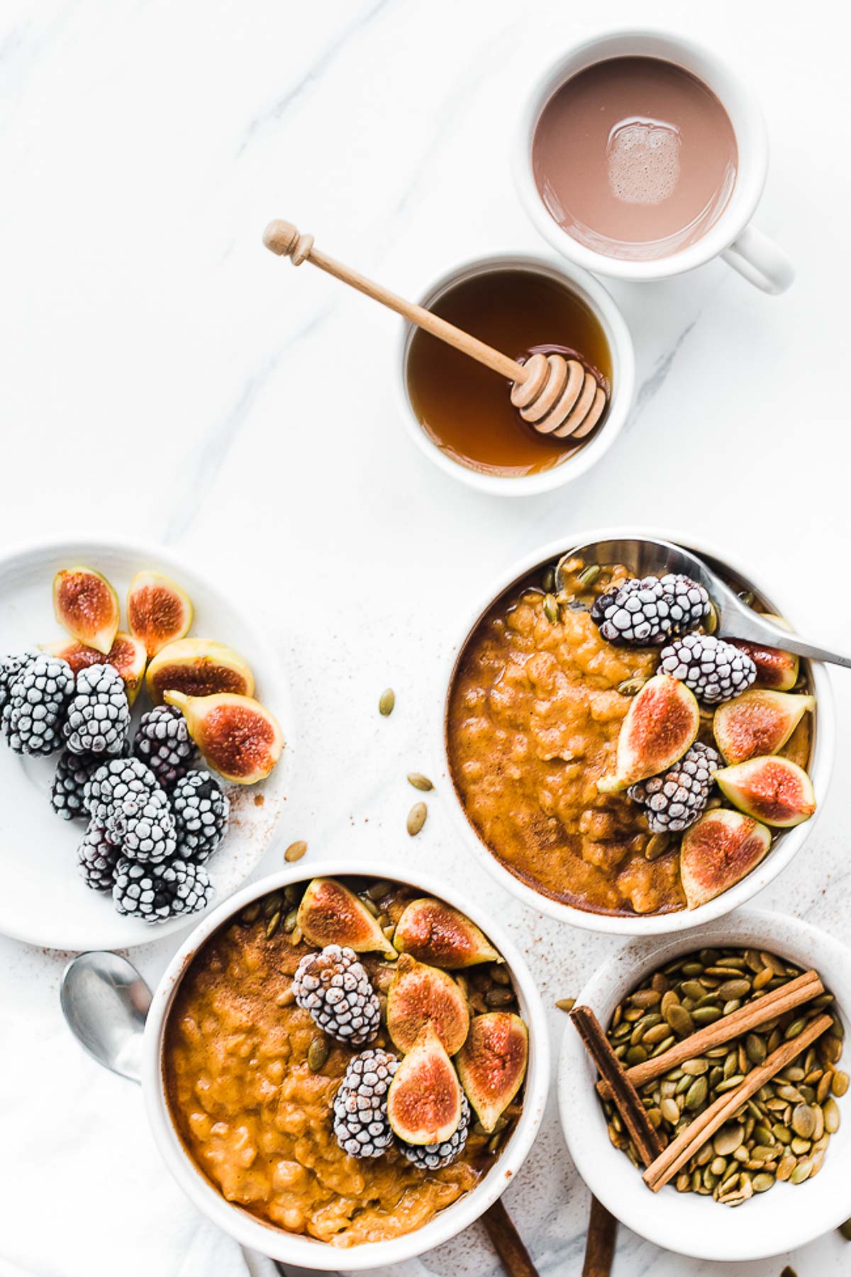 Two bowls of pumpkin oatmeal. The oats are garnished with berries and gas, and there are small bowls of berries and honey to the side.