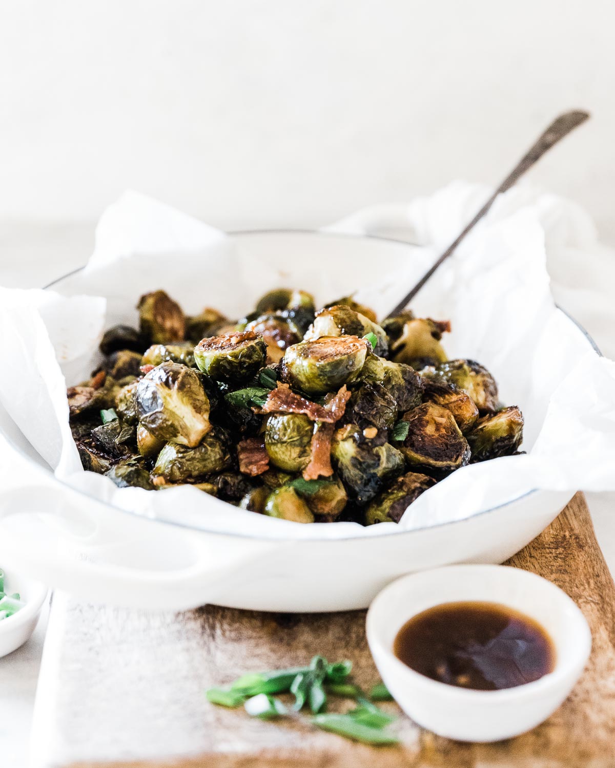 A ¾ view of roasted Brussels sprouts in a white braiser. There is a bowl of teriyaki to the side.