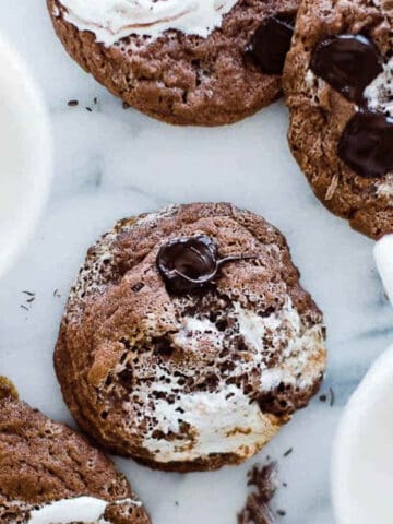 Chocolate marshmallow cookies directly on a surface with mugs of milk on the table too.