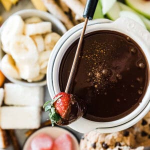 Chocolate Fondue in a white fondue pot surrounded by fruit.
