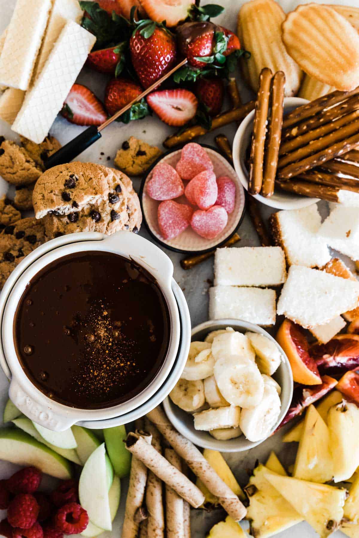 Chocolate fondue on a marble tray surrounded by apples, pretzels, pineapple, and pound cake.