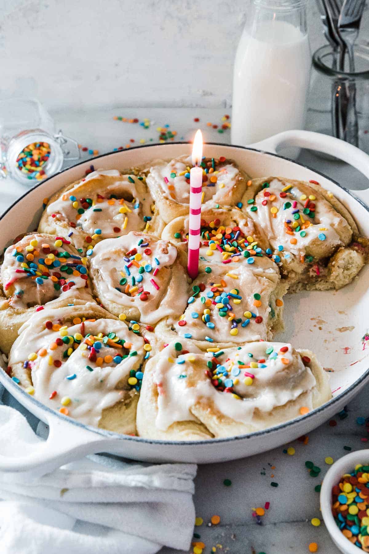 Birthday cake cinnamon rolls in a white braiser. There is a lit candle in the middle and the rolls are garnished with sprinkles.
