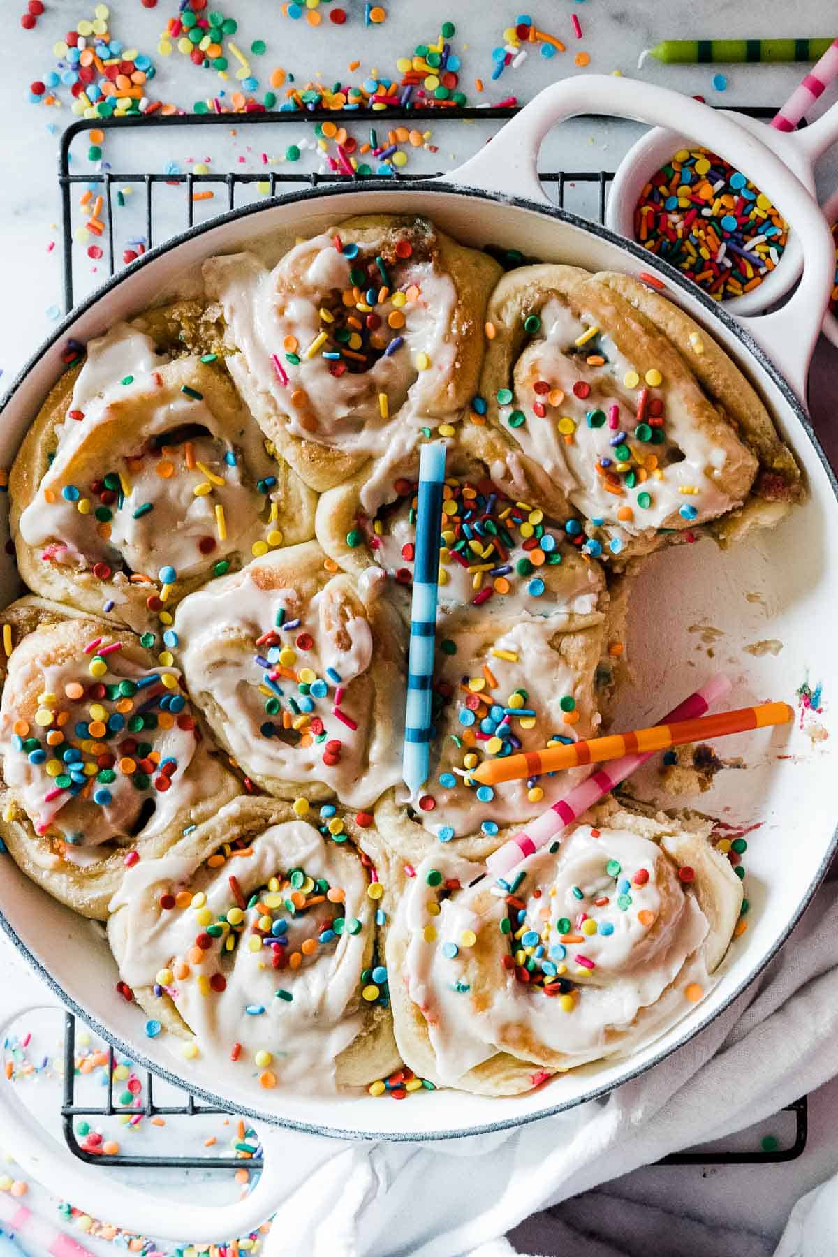 Birthday cake cinnamon rolls in a white braiser. They are topped with sprinkles and birthday candles.