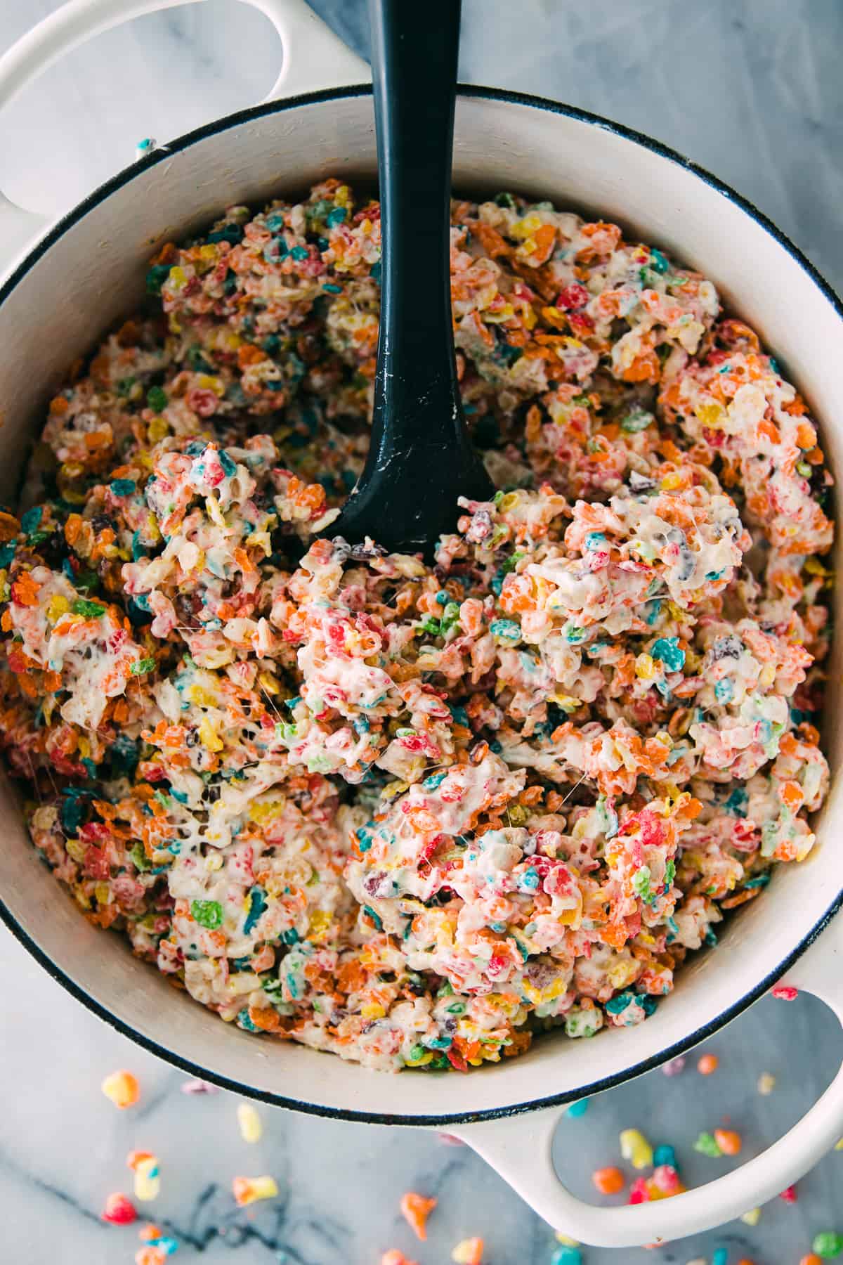 rainbow colored fruity pebbles stirred into marshmallows in a large white cast iron pan. 