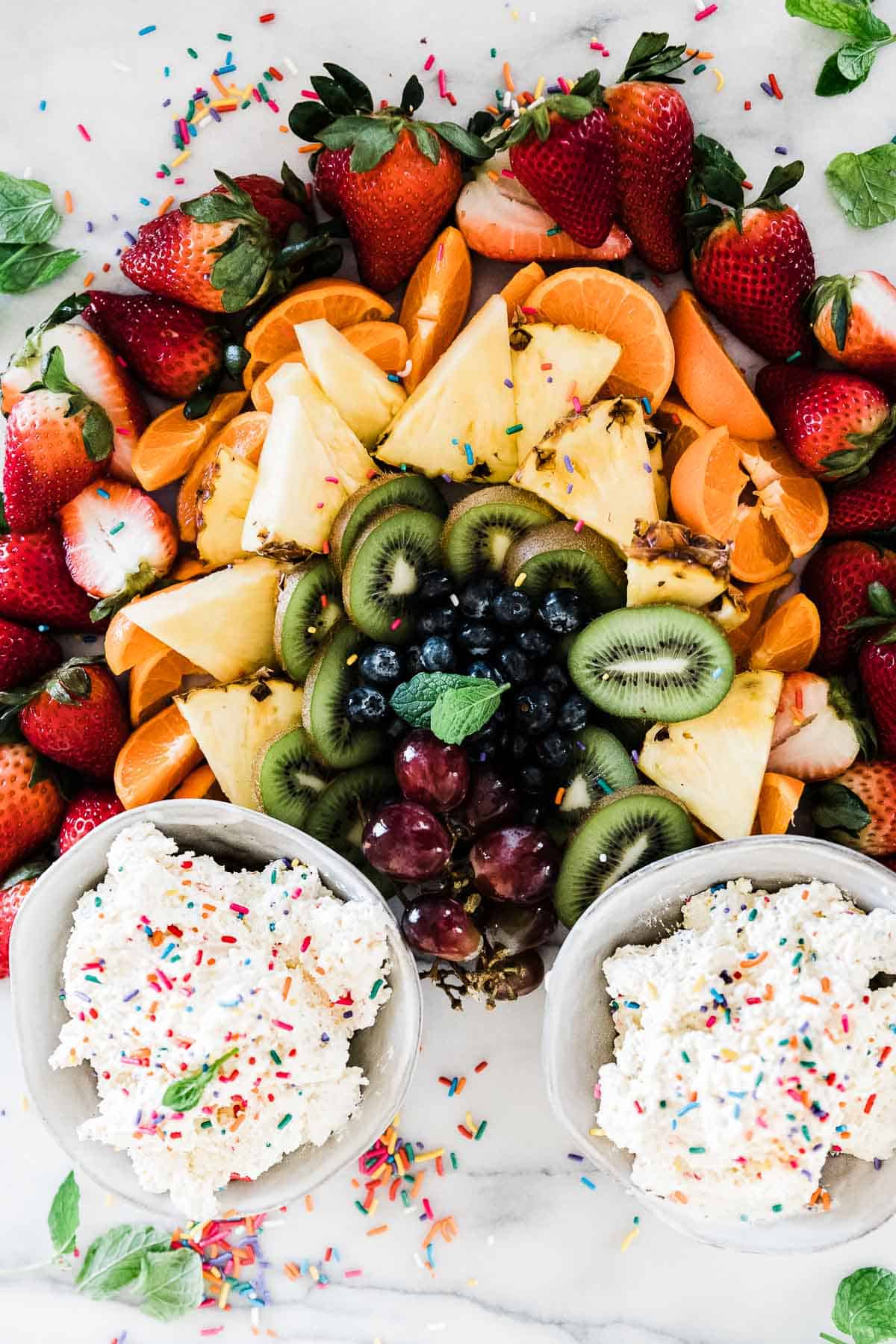 Cake batter dip in two bowls surrounded by fruit arranged into a rainbow.