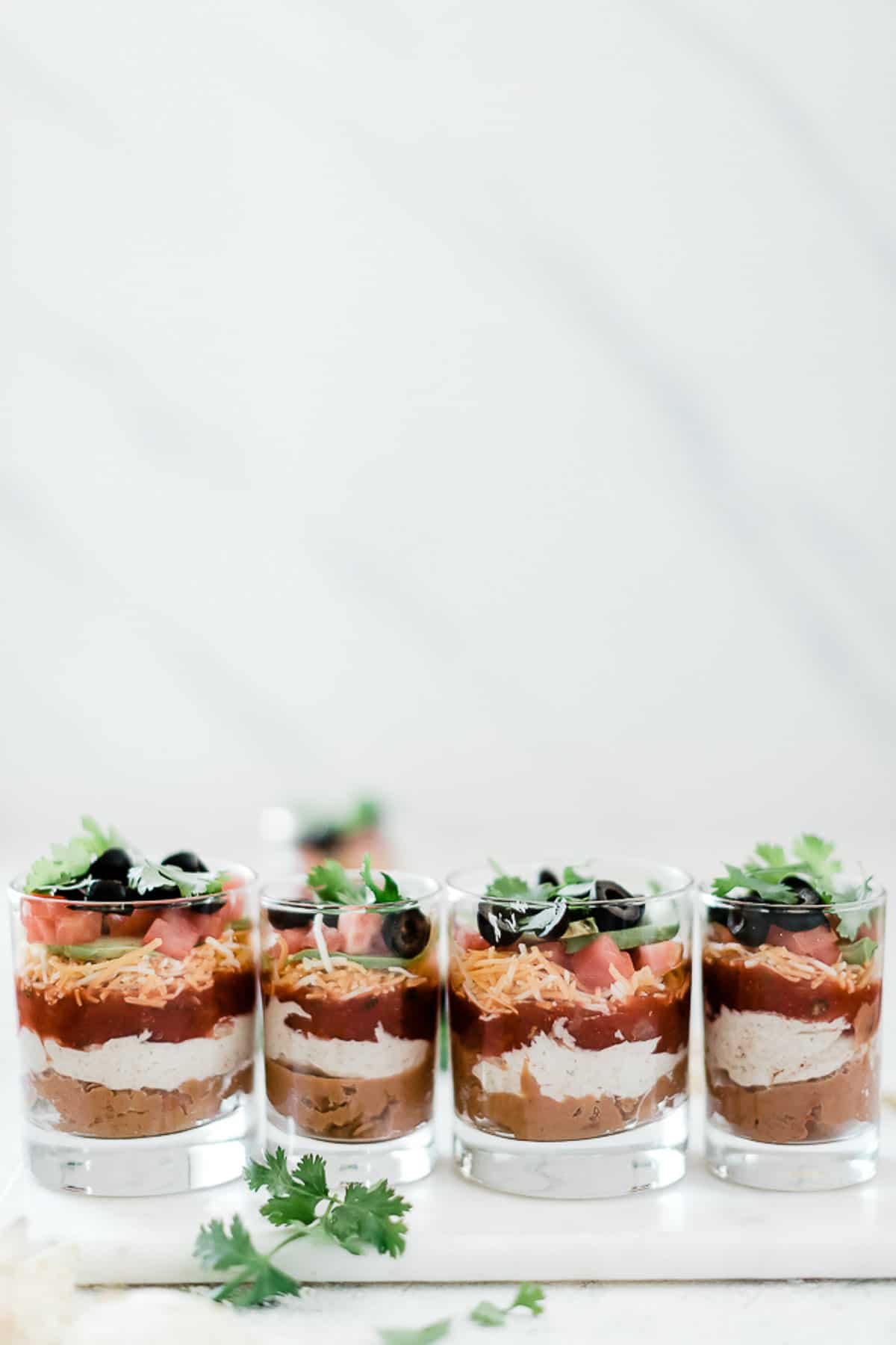 4 individual layered taco dips lined up. There are variations in the size of the glasses.
