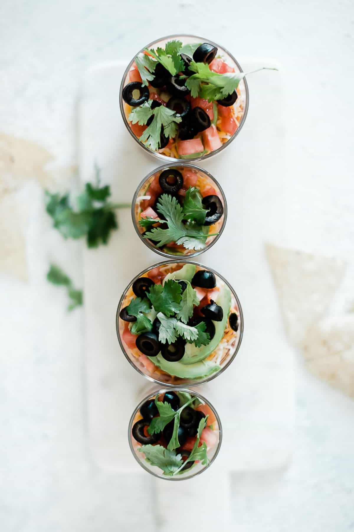 4 mini layered taco dips lined up on a marble tray. There are garnished with cilantro.