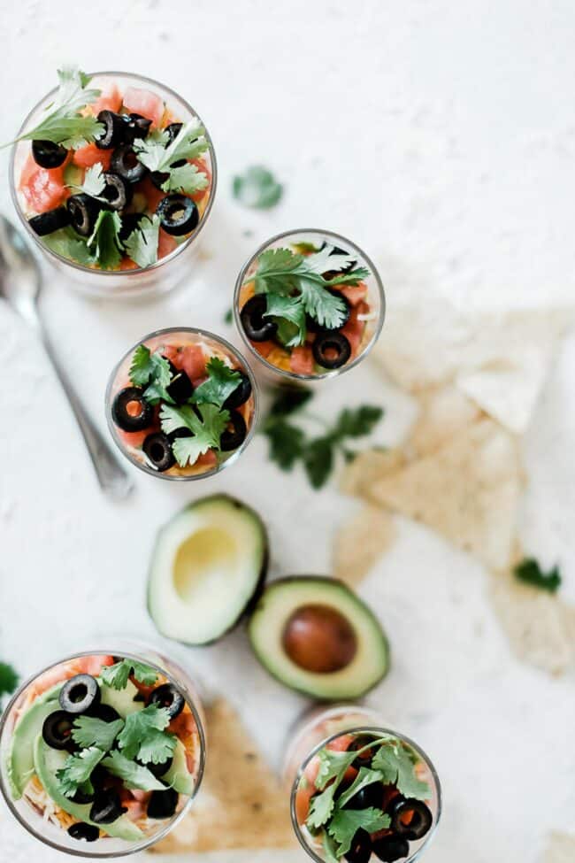 An overhead shot of layered taco dip cups on a white counter. There are chips and avocado to the side.