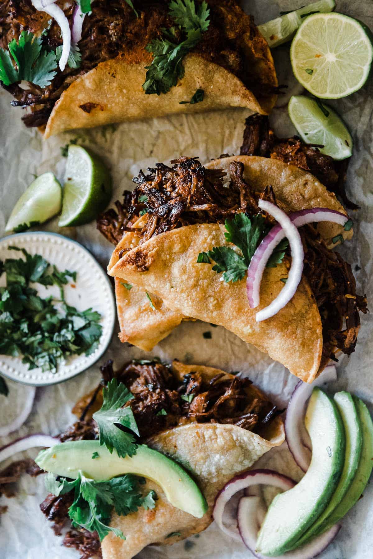 Instant pot barbacoa beef tacos on parchment paper. They are garnished with avocado and red onion.