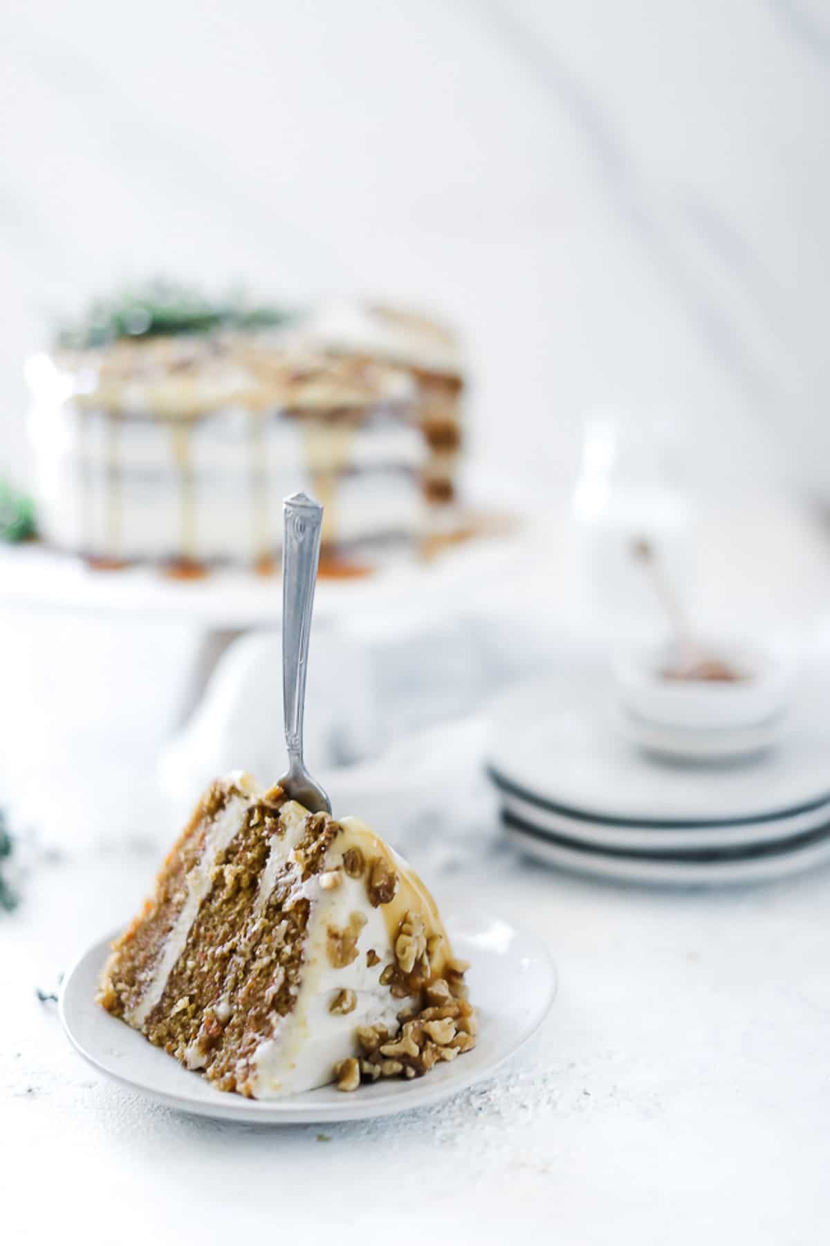 A slice of carrot cake on a white plate with a fork stuck in it. There is a full cake in the background.