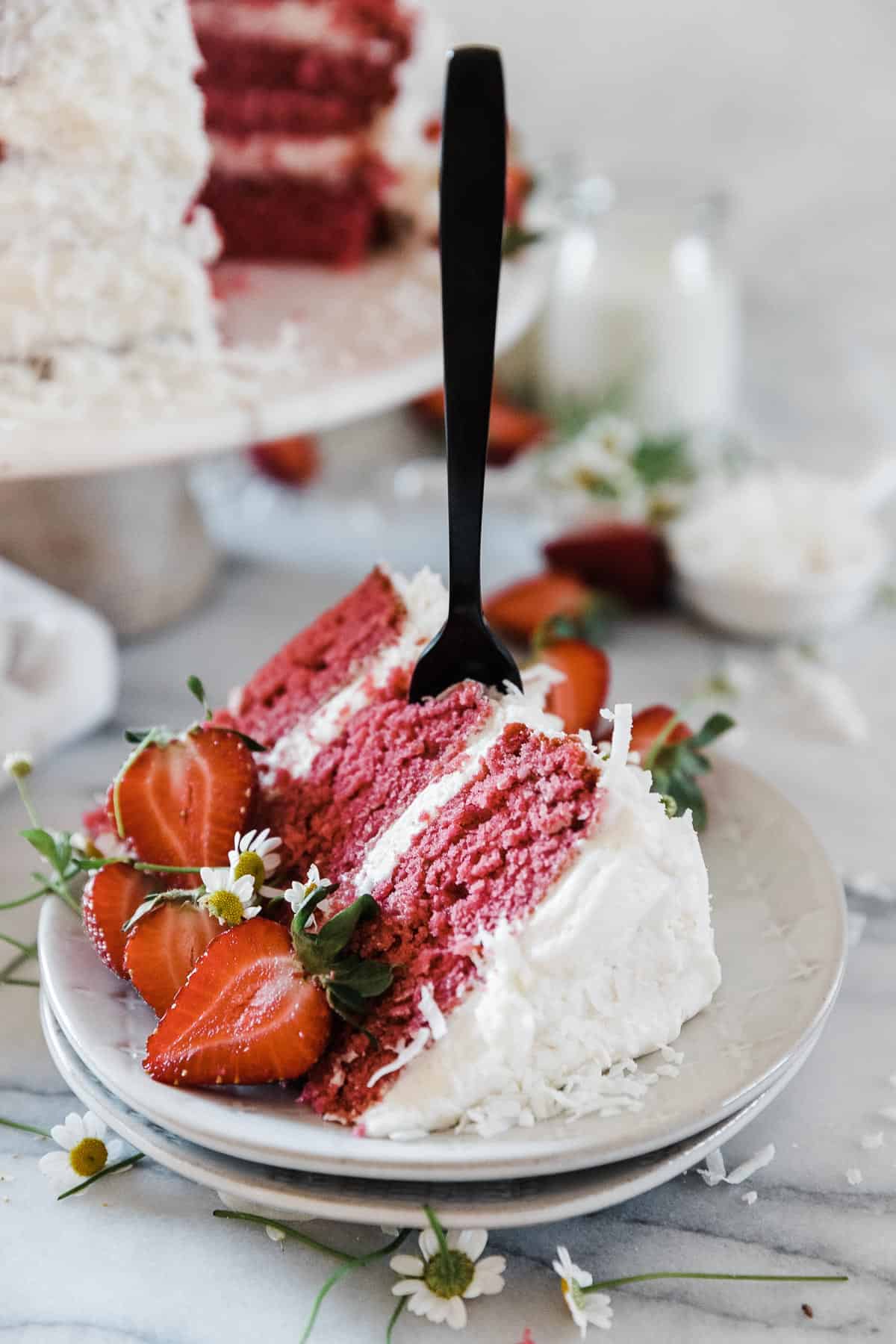 A large slice of strawberry coconut cake on a white dessert plate. There is a black fork stuck into the slice.