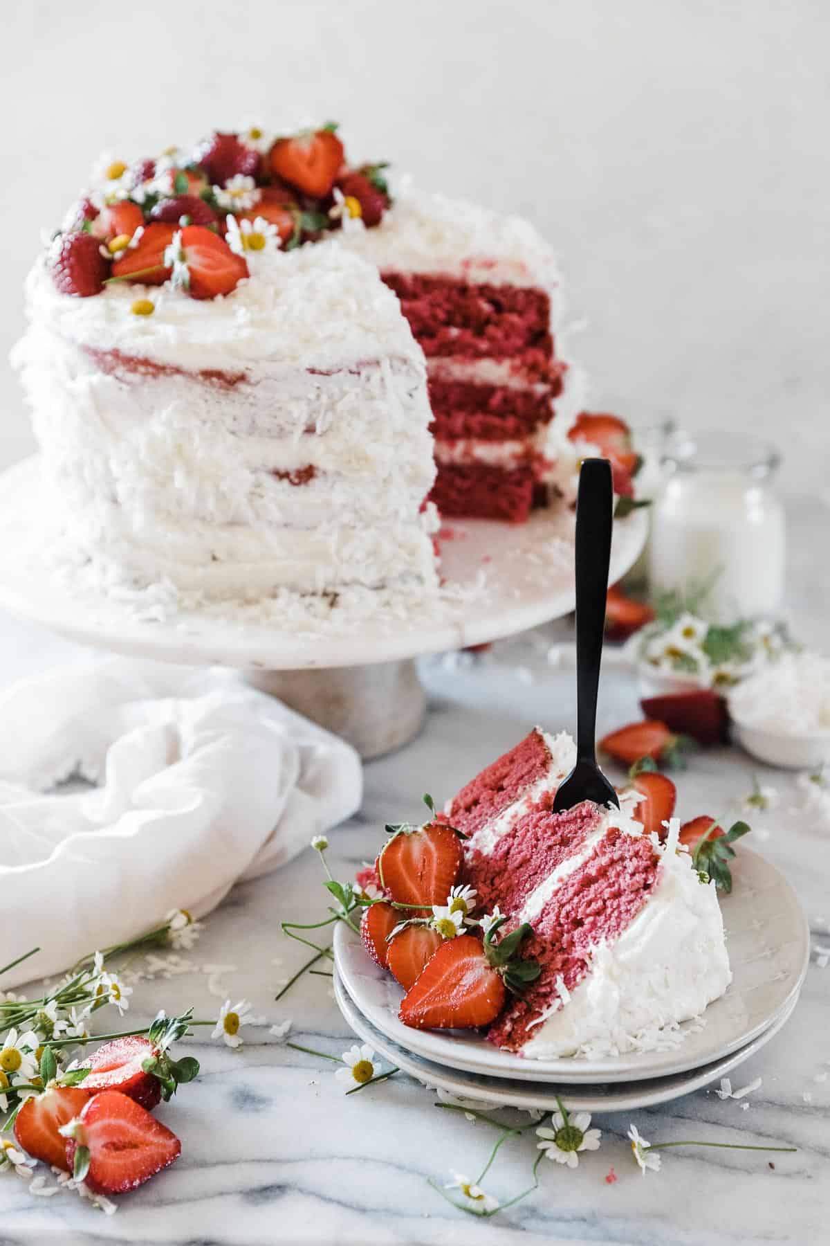 A slice of strawberry coconut cake on a small plate, with a full cake in the background.