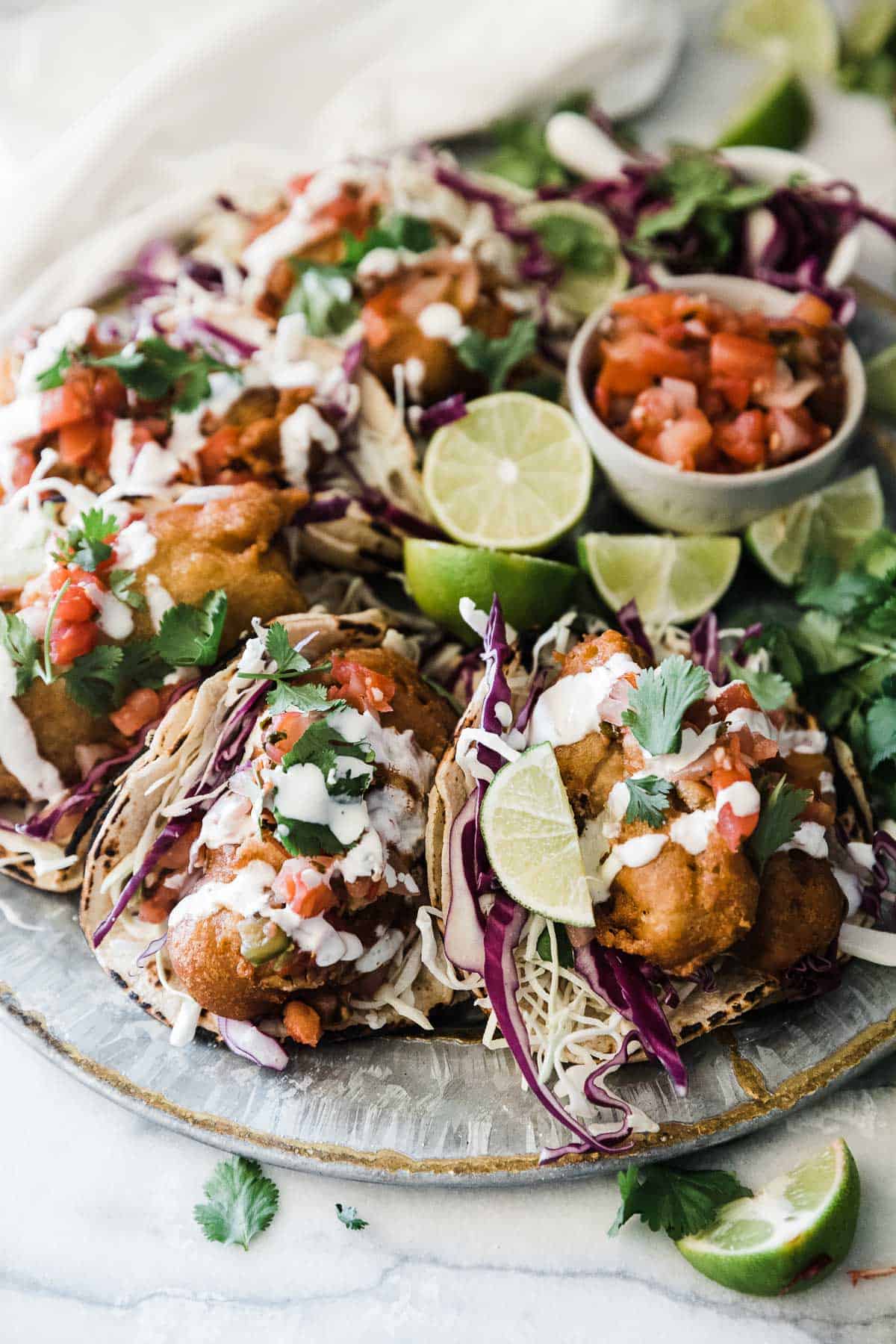 A ¾ view of beer battered fish tacos on a round silver platter. They are garnished with cilantro and pico.