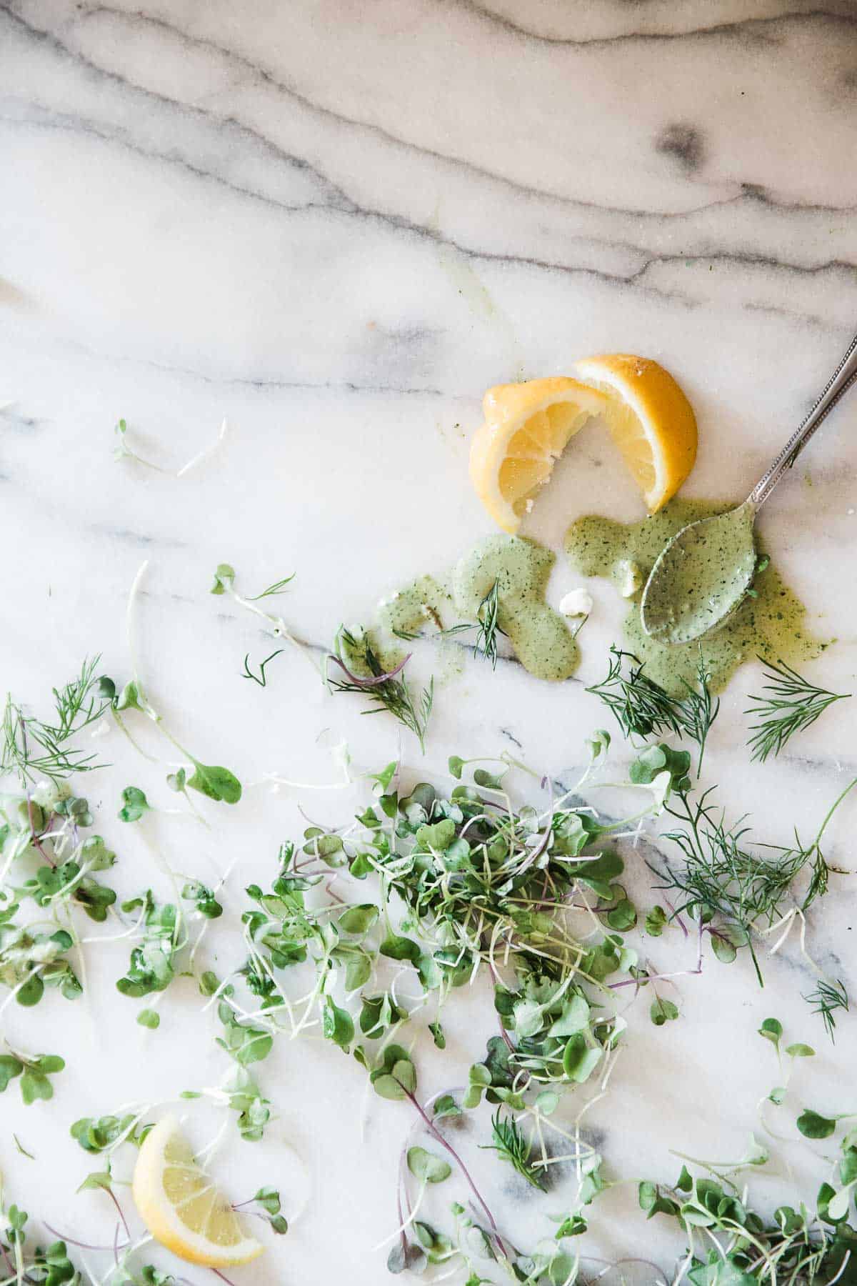 A spoon full of green goddess dressing on a marble counter surrounded by lemons and micro greens.