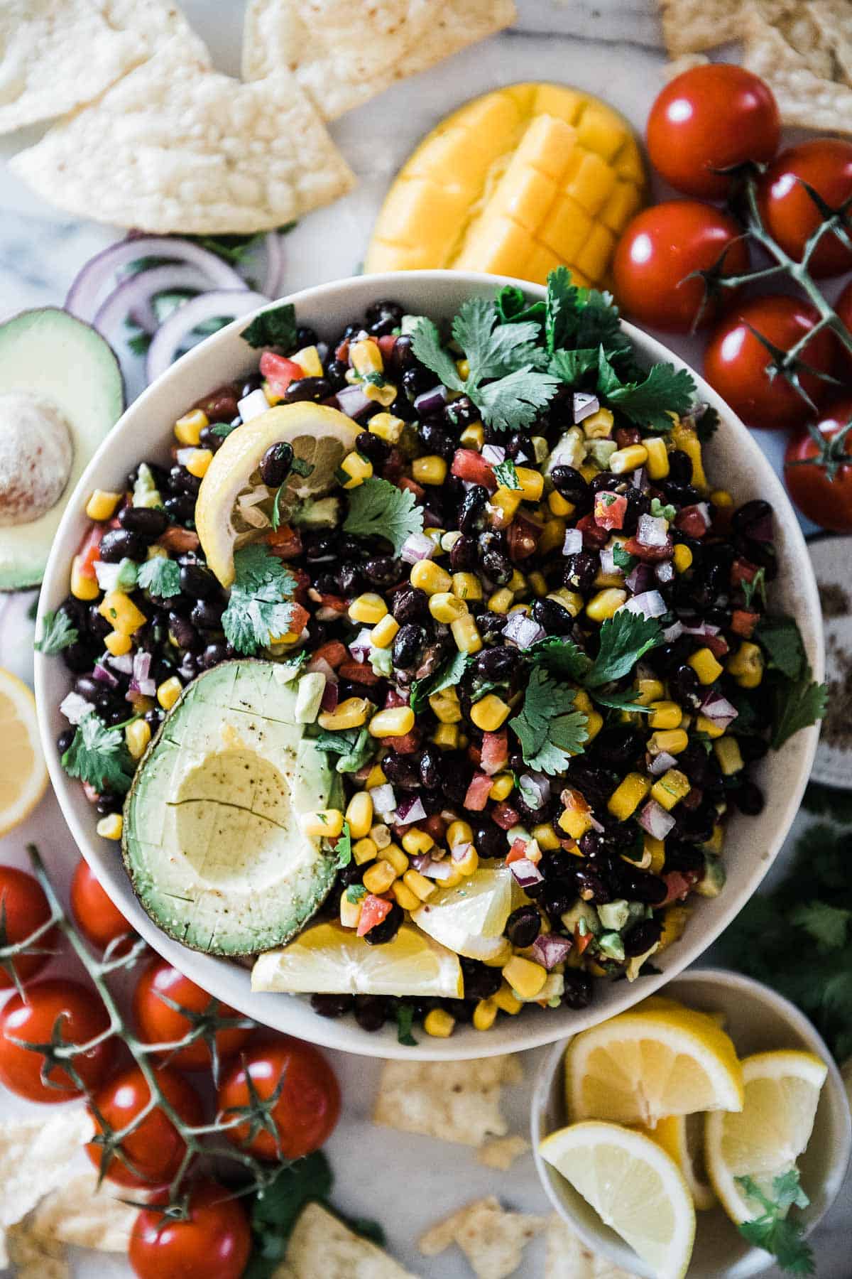 Black bean salsa in a large while bowl. It is garnished with limes, avocado, and lemons.