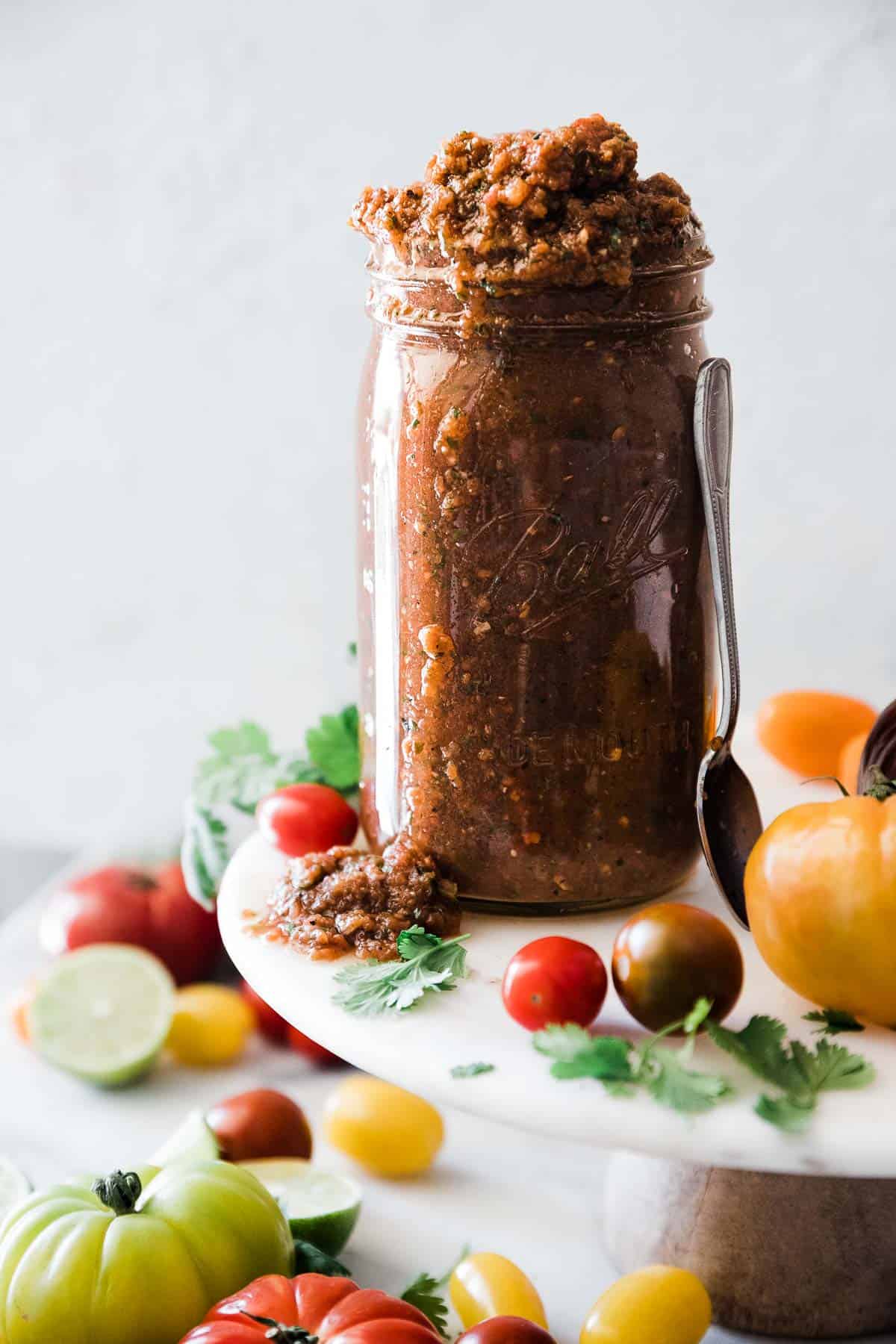 Chipotle salsa in a mason jar atop a cake stand. There are tomatoes and limes around it.