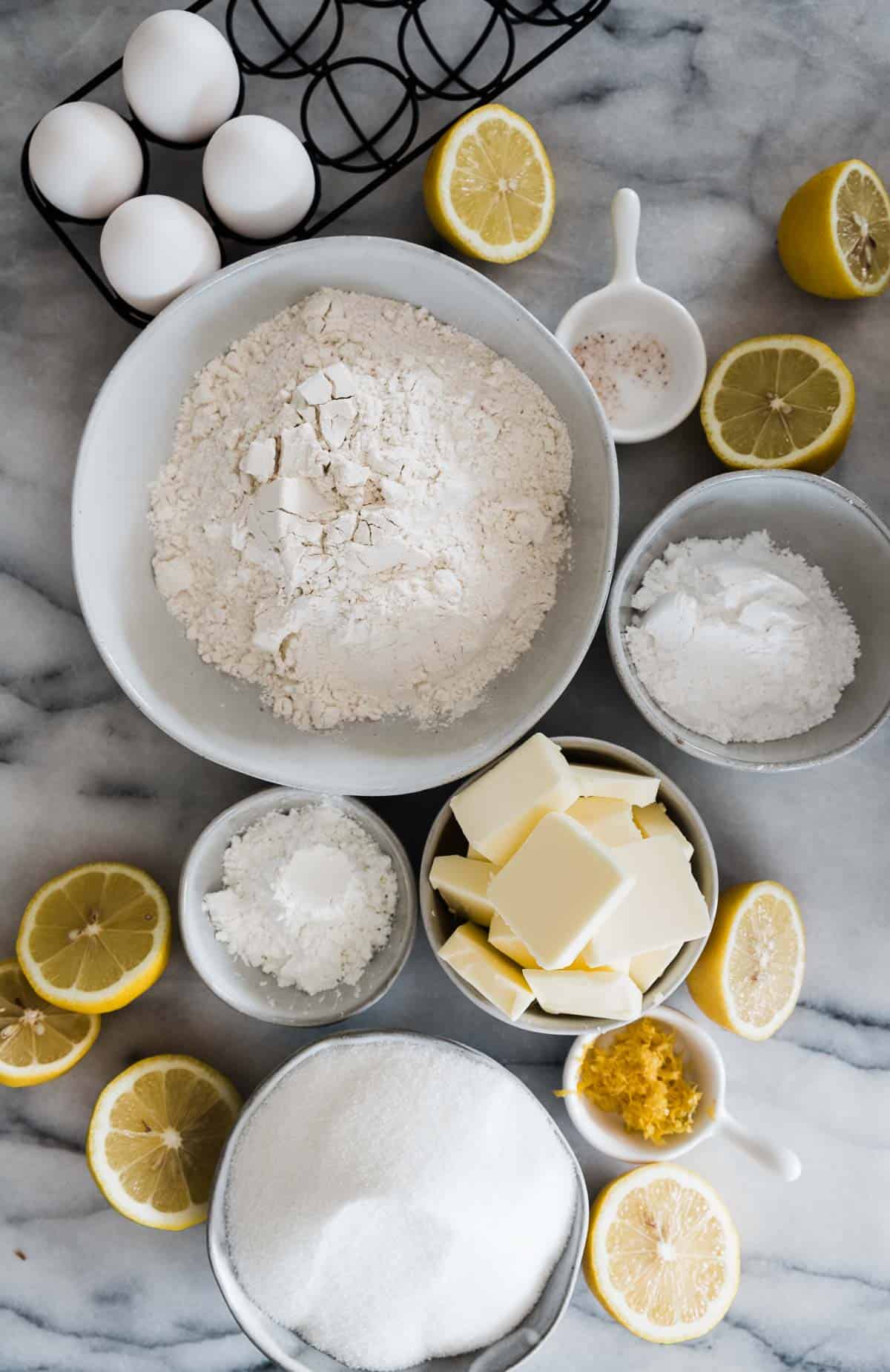 Ingredients needed to make lemon bars placed in bowls and set on a marble counter.