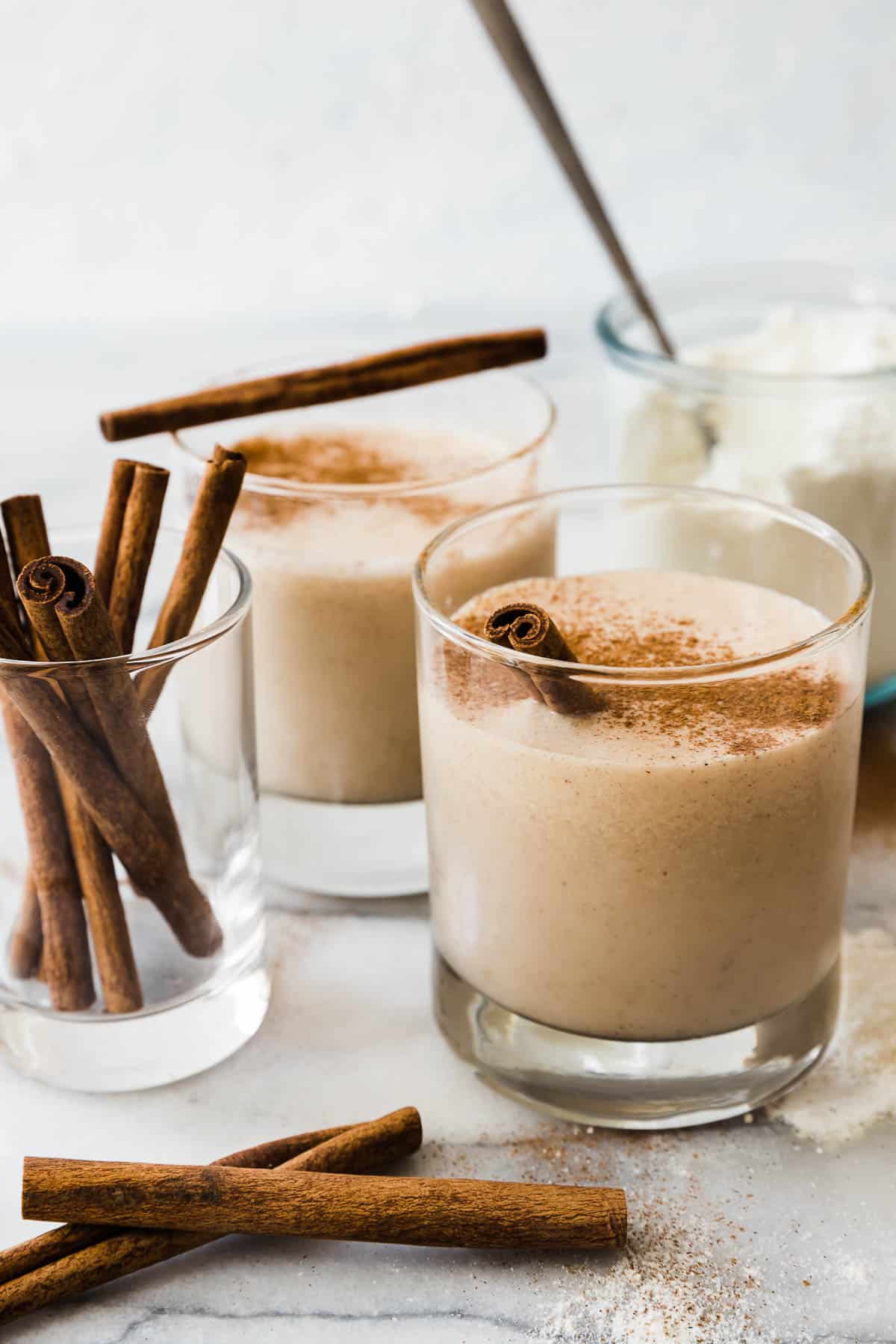 Two glasses of vanilla protein shake set beside a glass of cinnamon sticks. There is a jar of vanilla protein powder in the background.