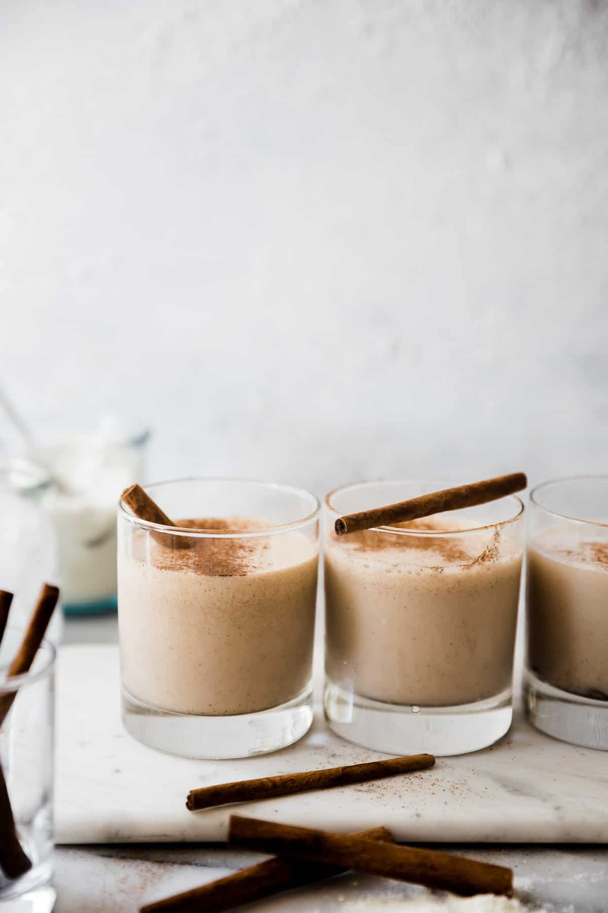 Three glasses of snickerdoodle protein shake lined up on a marble tray. They are garnished with cinnamon sticks.
