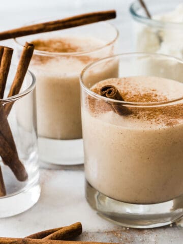 A close up of snickerdoodle protein shake in a glass. There are cinnamon sticks to the side.