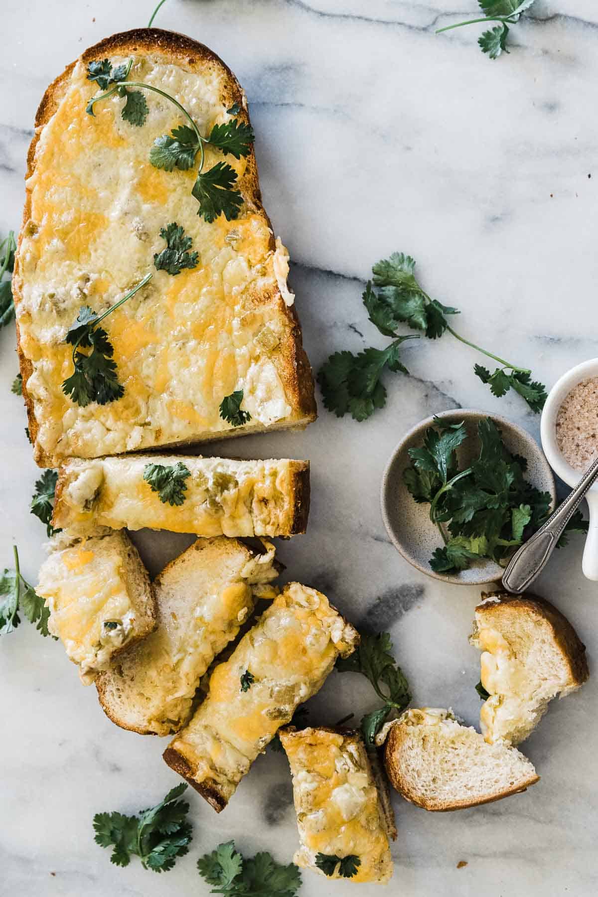 Chili cheese toast sliced and laid on a marble counter. There are small bowls of salt and cilantro to the side.