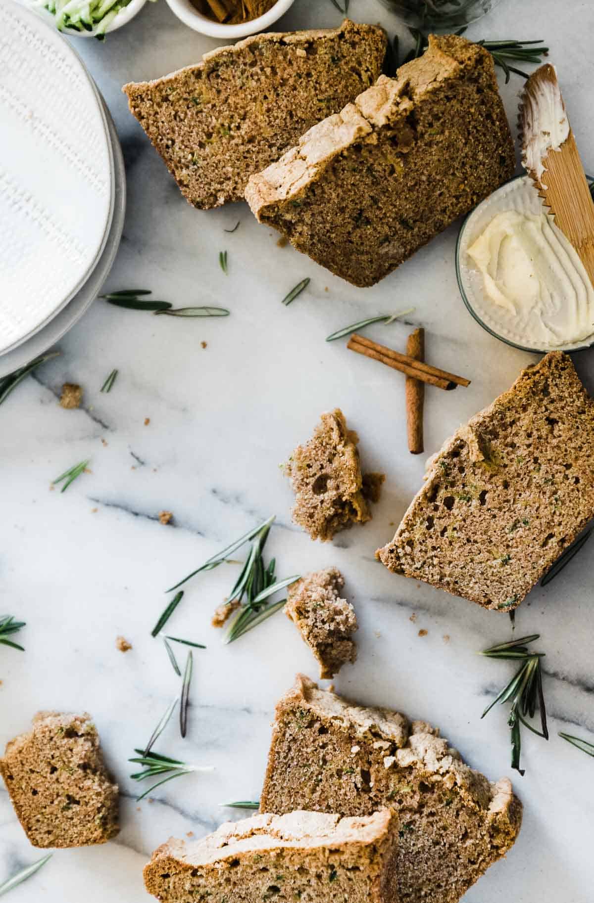 Slices of easy zucchini bread recipe laid on a marble counter. There are crumbs, rosemary sprigs, and cinnamon scattered around.