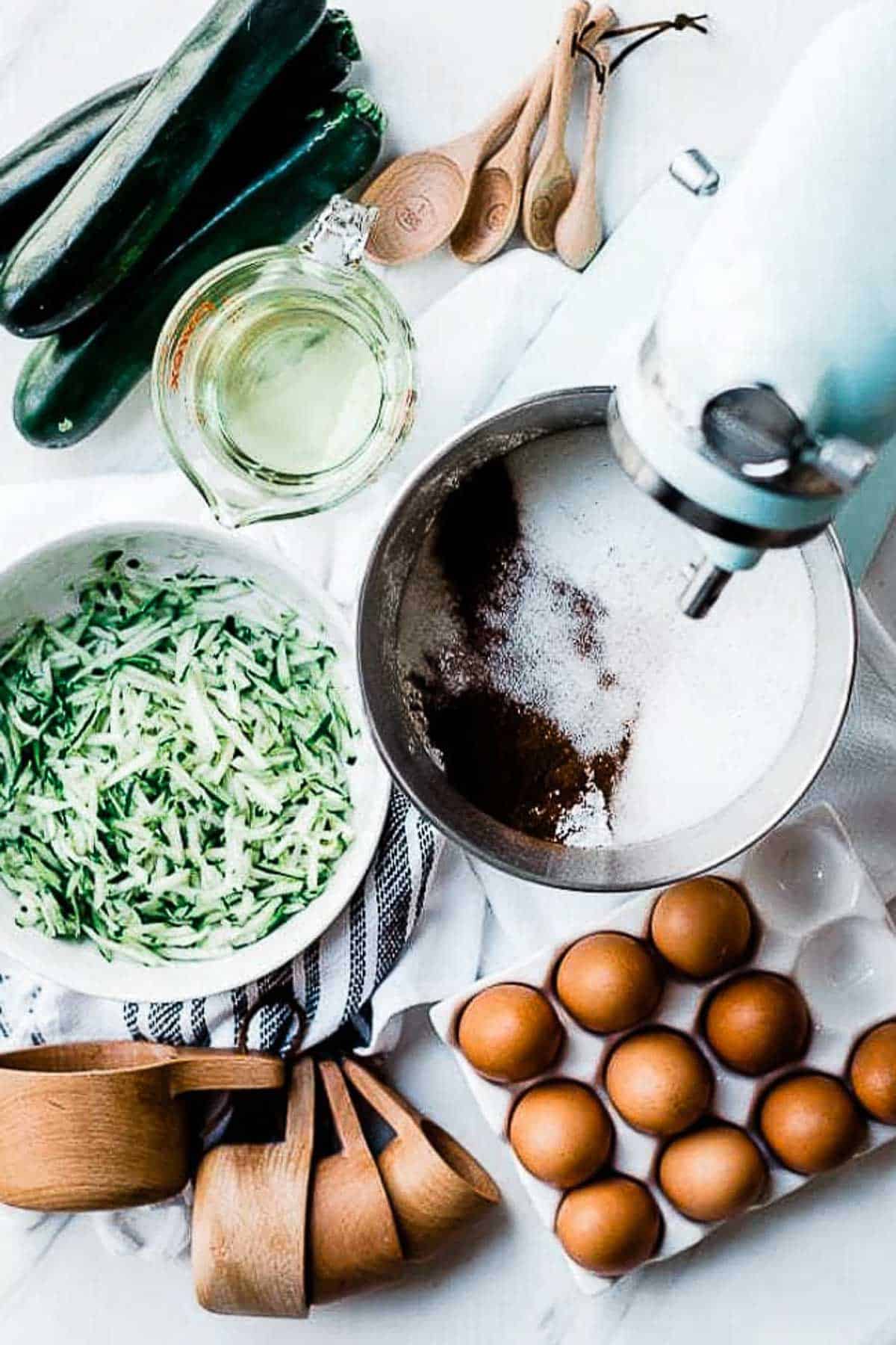 Zucchini, oil, eggs, measuring cups, and a bowl of flour and sugar on a white counter.