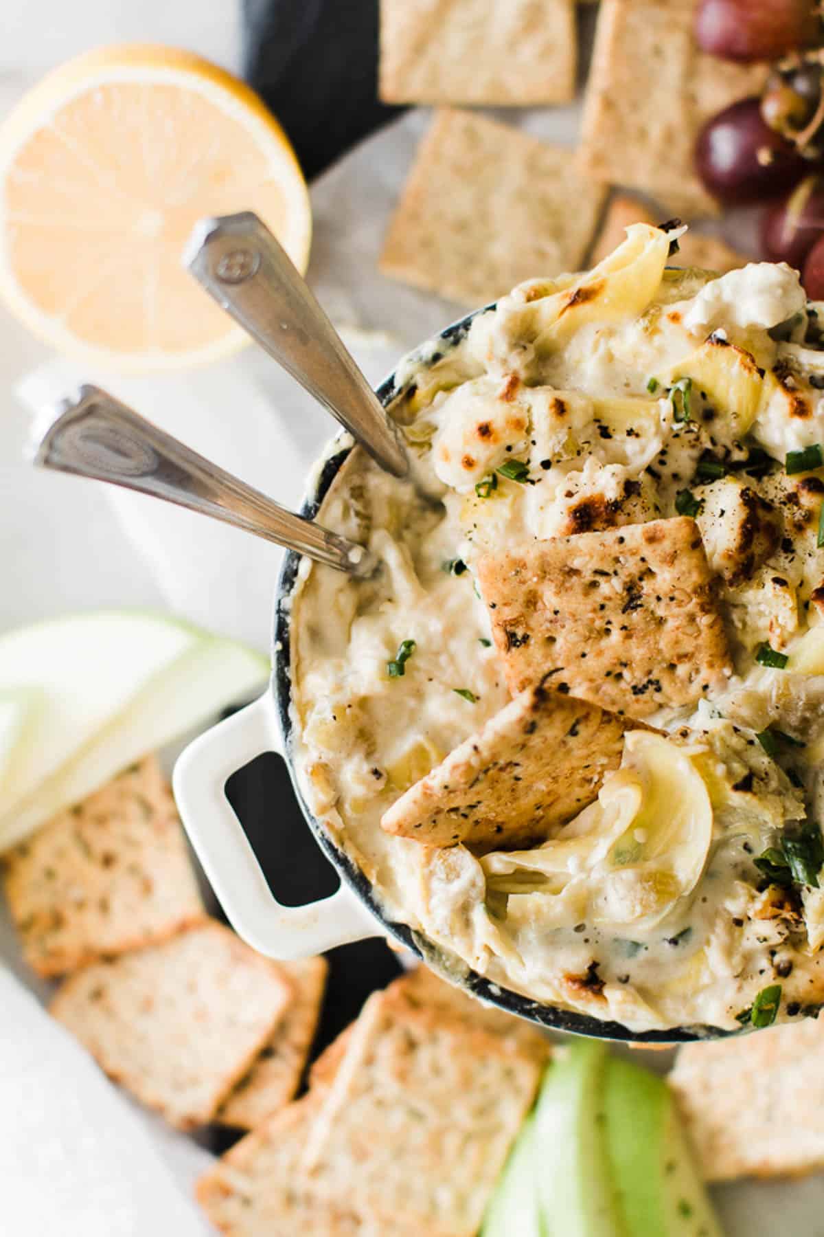Bowl of hot artichoke dip with two spoons and crackers served up on a table.